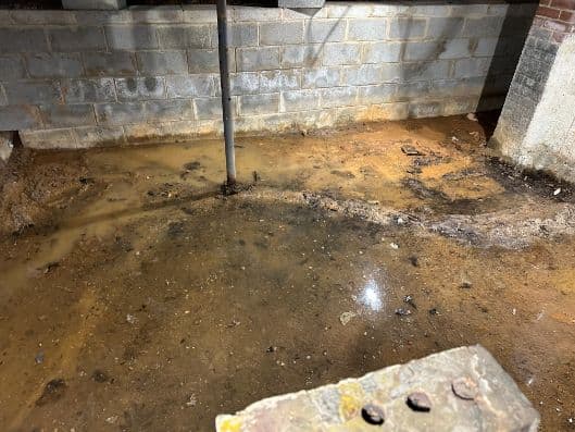 Muddy, waterlogged basement floor beneath a cinder-block wall, with a vertical pipe, puddles, debris, and a concrete block in front.
