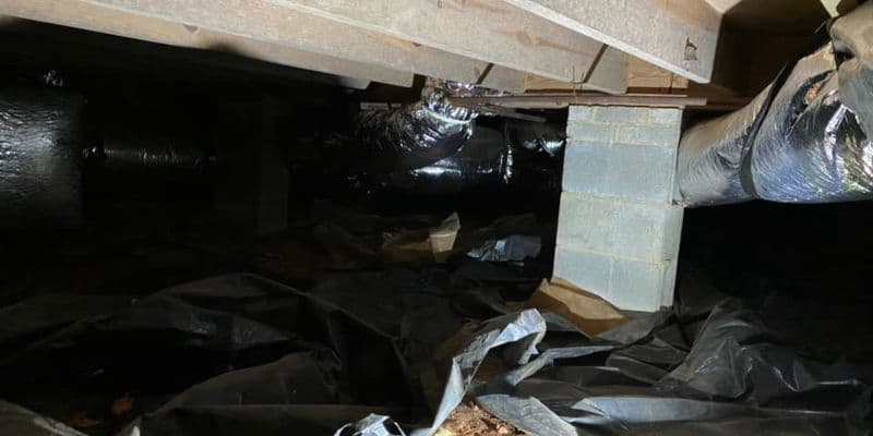 Dark crawlspace beneath a building with black plastic sheeting, exposed beams, a brick support, and wrapped HVAC ducts, conveying neglect.