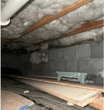 Dim crawlspace with exposed gray insulation, wooden ceiling joists, cinder block wall, scattered boards, debris, and a cramped, unfinished atmosphere.