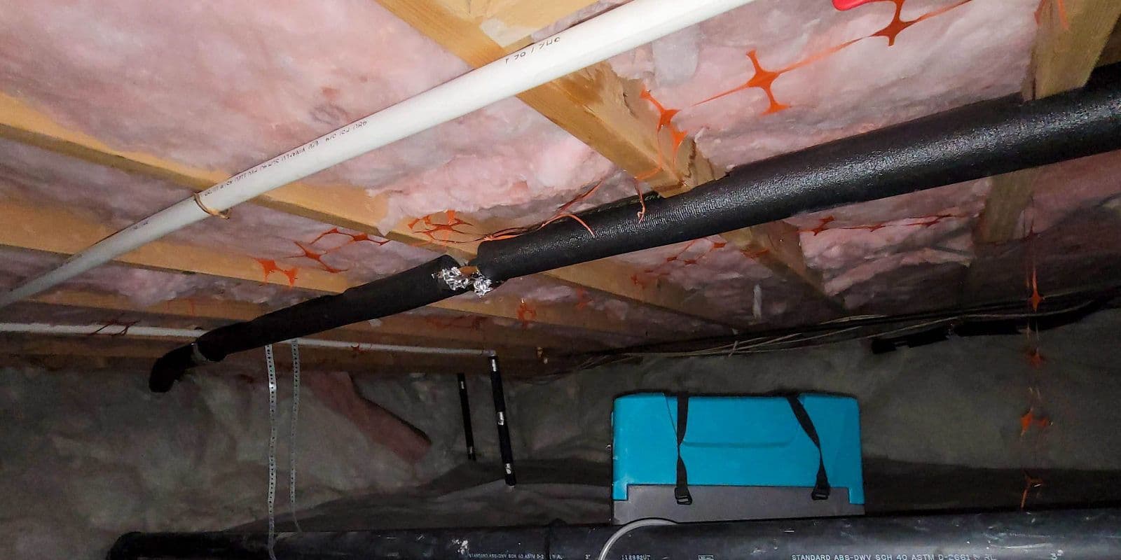 Low-ceiling crawlspace with exposed pink insulation, wooden beams, black and white utility pipes, and a bright blue panel in the background.