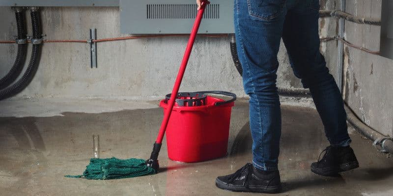 Person in jeans mops a wet concrete floor with a red mop and bucket beside a green mop head in an industrial utility room.