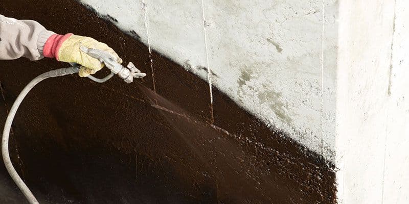 Gloved worker sprays a dark protective coating onto a concrete wall with a pressurized hose, creating a diagonal black layer.