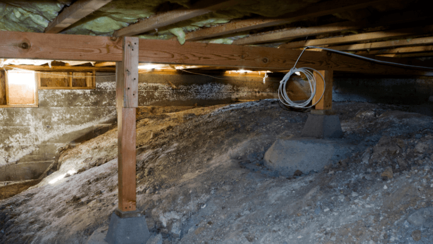 Damp, dim crawl space beneath a house with wooden support beams, concrete footings, exposed insulation, uneven soil, and coiled wiring.