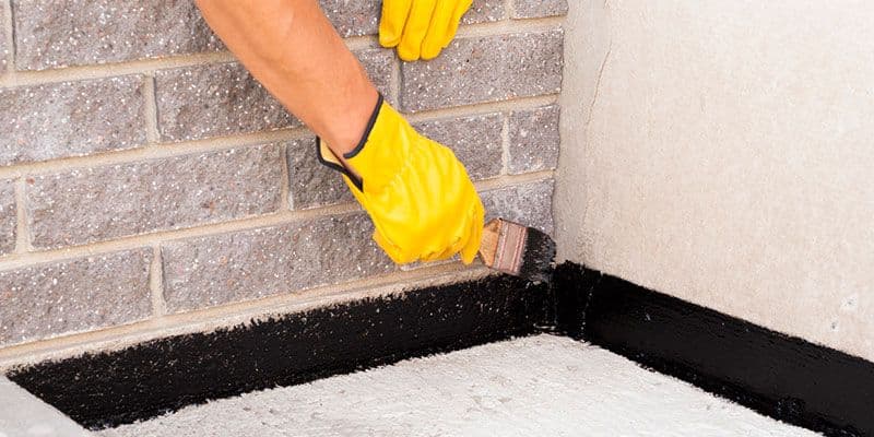 Worker in yellow gloves applies black waterproofing sealant with a brush along the base of a gray brick wall and concrete corner.