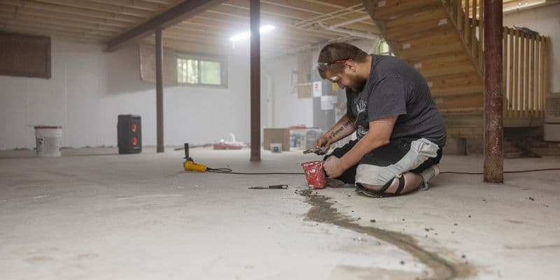 Worker kneels on a concrete basement floor using a red power tool, with scattered tools and exposed beams suggesting focused renovation work.