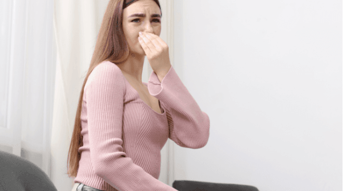 Worried young woman in a pink sweater covers her mouth with one hand, appearing nauseated or ill while seated indoors against a light wall.