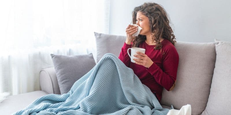 Woman in a red sweater sits on a sofa wrapped in a blue blanket, holding tea and tissues, suggesting a cozy moment while feeling unwell.