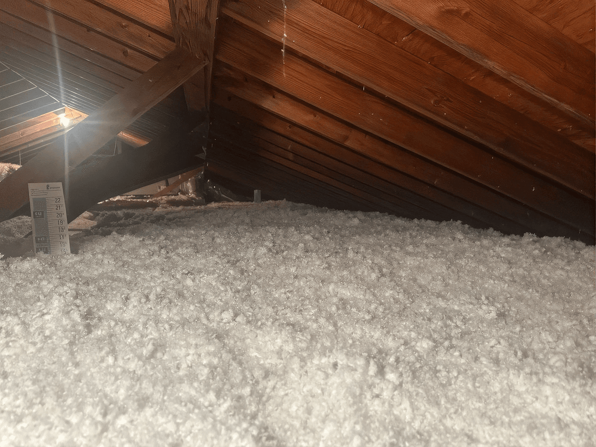 Attic with freshly installed blown-in insulation covering the attic floor between exposed wooden roof rafters, with an insulation depth ruler visible to verify proper coverage and energy efficiency.