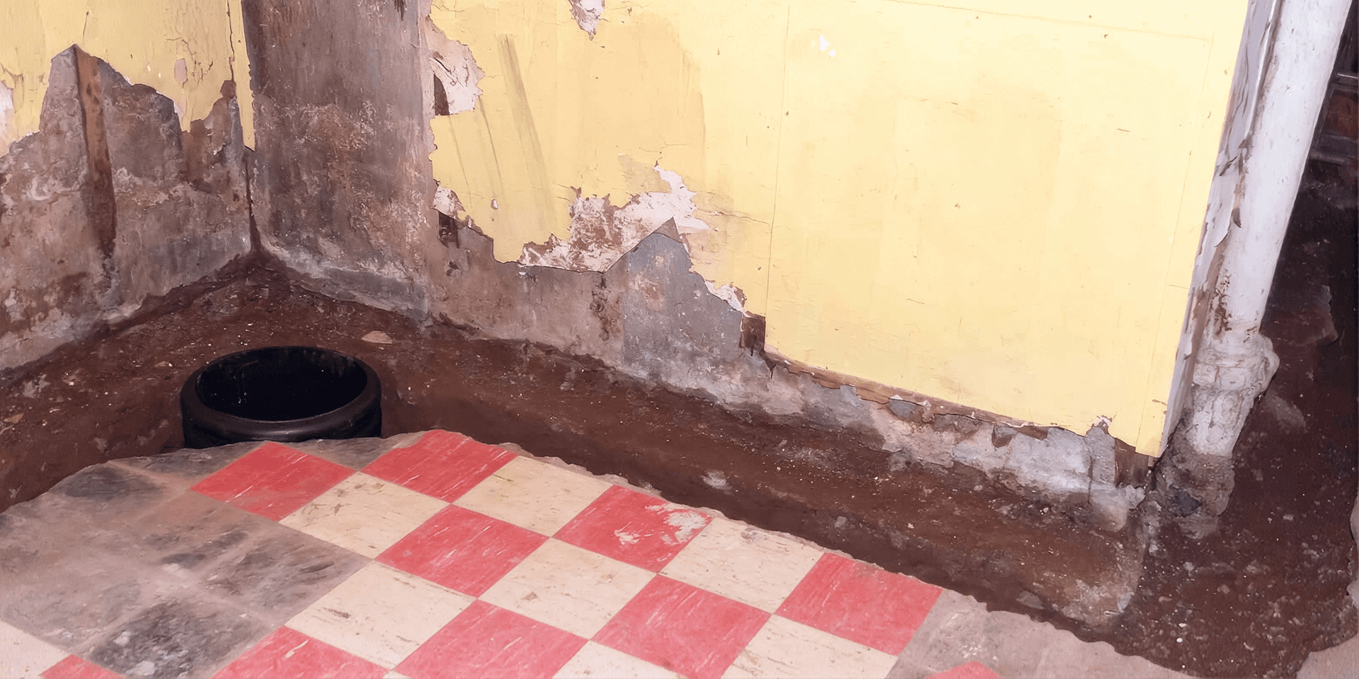 Basement with exposed soil around the foundation walls, peeling paint, moisture damage, and an open drainage or sump pit during waterproofing work.