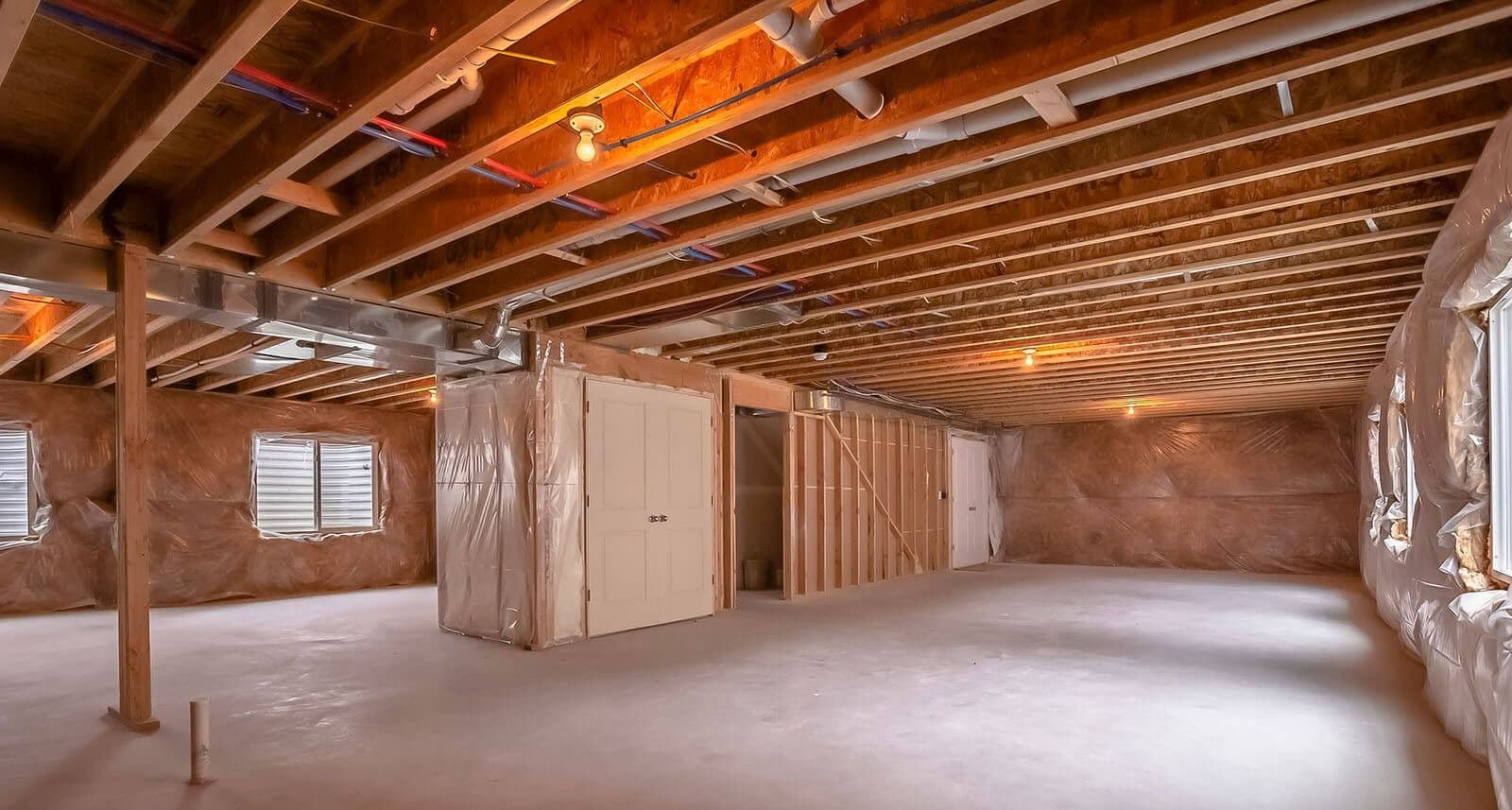 Unfinished basement with exposed ceiling joists, concrete floors, framed utility rooms, plastic moisture barriers along the walls, support posts, and overhead plumbing and ductwork, providing a clean, open space ready for finishing or storage.