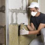 Thumbnail image of Technician wearing a face mask and gloves installing fiberglass batt insulation inside an unfinished wall cavity, improving the home's energy efficiency and thermal performance.