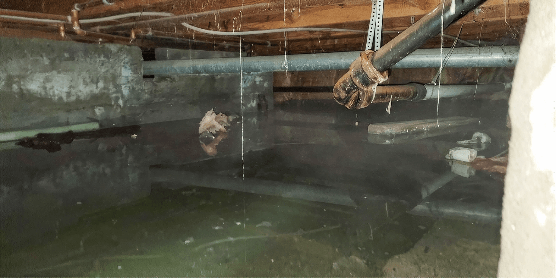 Flooded crawl space with standing water beneath exposed plumbing and floor joists, highlighting severe moisture intrusion that can lead to structural damage, mold growth, and poor indoor air quality.