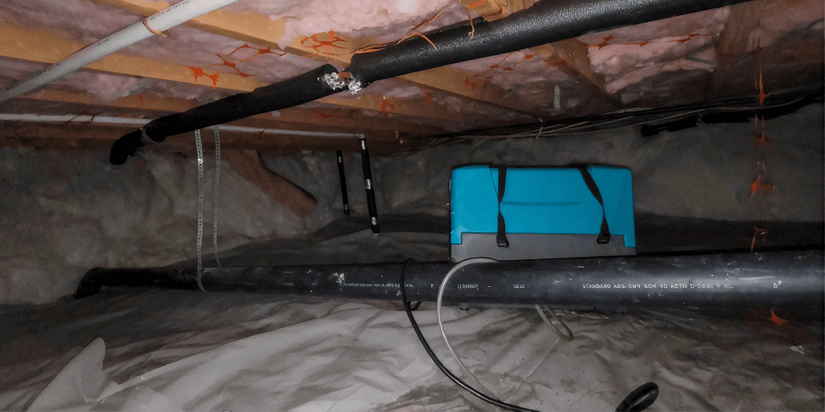 Crawl space with fiberglass insulation installed between floor joists, exposed plumbing and insulated piping, a crawl space dehumidifier, and a vapor barrier covering the ground to help control moisture and improve energy efficiency.