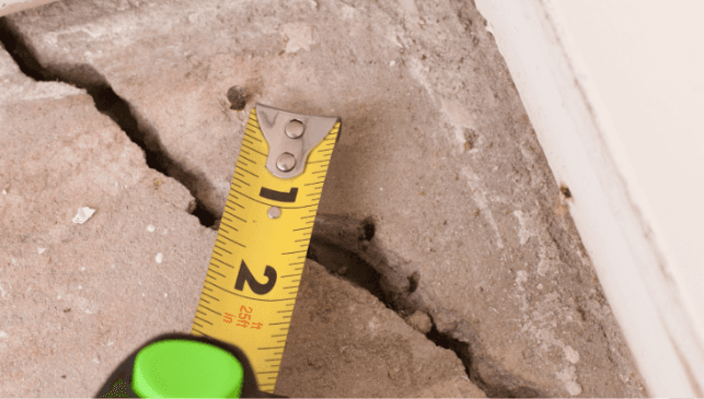 Close-up of a wide, jagged crack in a concrete surface measured with a yellow tape measure, highlighting structural damage and concern.