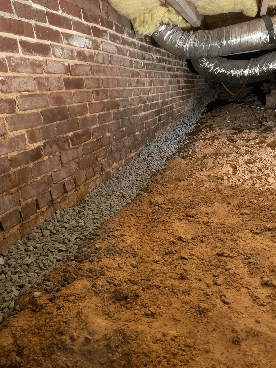Crawl space with exposed soil flooring, a brick foundation wall, overhead ductwork and insulation, and a newly installed perimeter drainage trench filled with gravel to help collect and redirect groundwater away from the home's foundation.